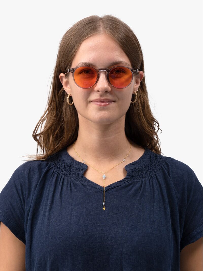 Frau in dunkelblauer Bluse mit runder Brille mit orangefarbenen Gläsern und grauer Fassung, goldenen Creolen und feiner Goldkette