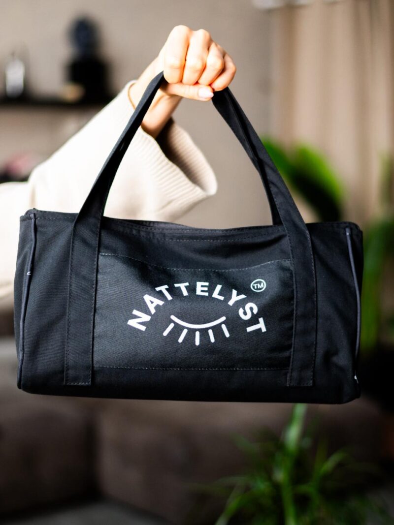 Schwarze Stofftasche mit NATTELYST-Logo an einem Henkel gehalten, Nahaufnahme vor unscharfem Wohnzimmerhintergrund