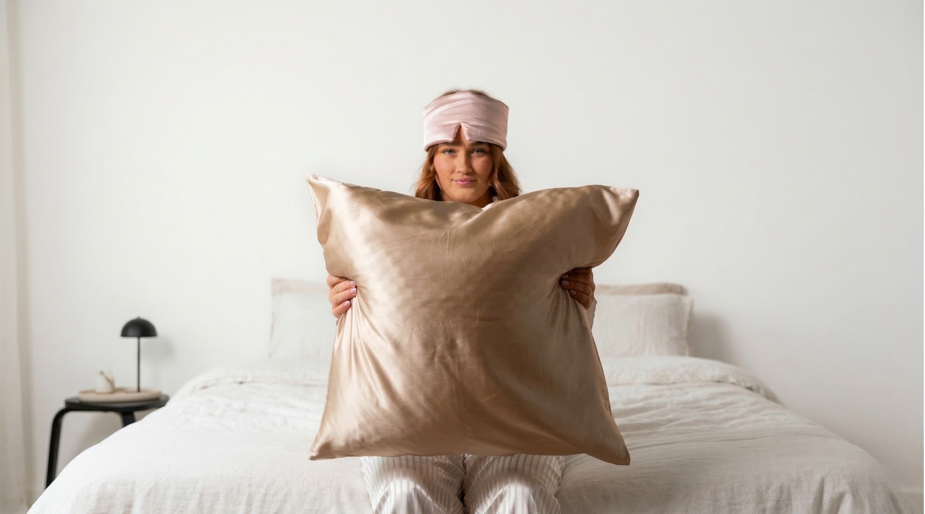 Frau mit Seiden-Schlafmaske hält ein großes champagnerfarbenes Seidenkissen in einem minimalistischen Schlafzimmer