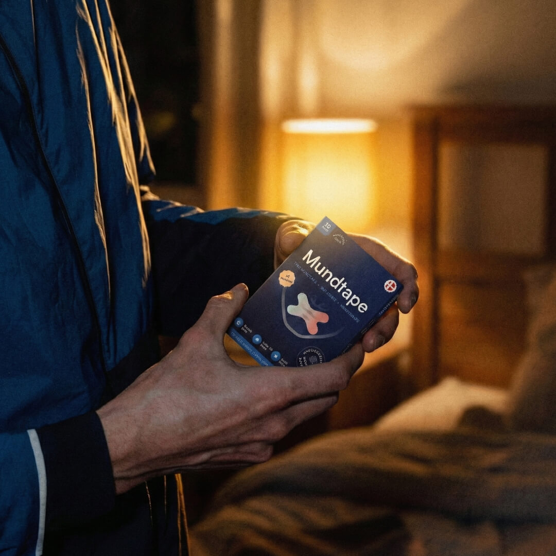 Hände halten Packung Mundtape vor warm beleuchtetem Schlafzimmer, entspannte Abendstimmung