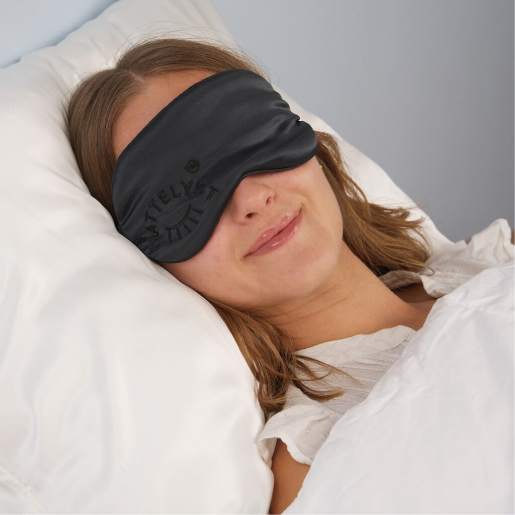 Frau liegt lächelnd im Bett mit schwarzer Schlafmaske auf weißem Kopfkissen