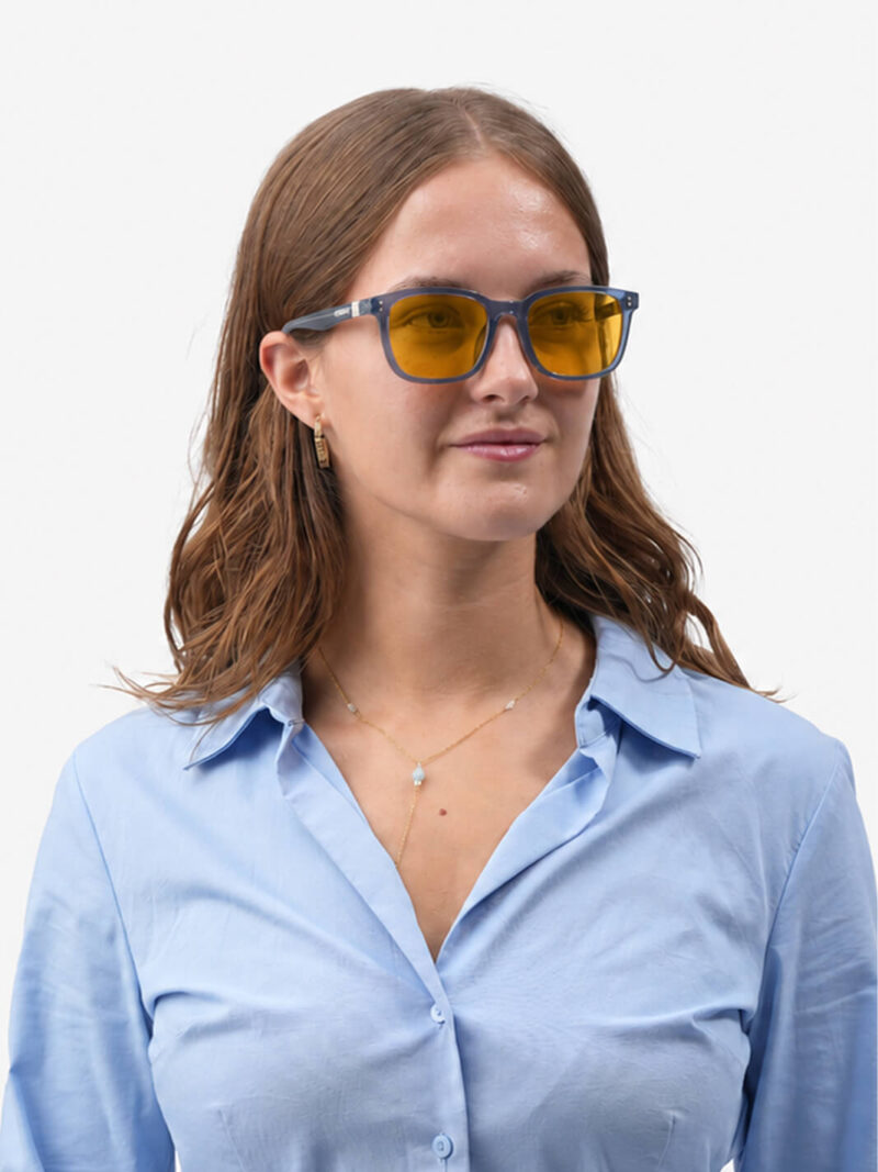Frau mit blauer Brillenfassung und gelben Gläsern, hellblaue Bluse, goldene Kette und Ohrringe