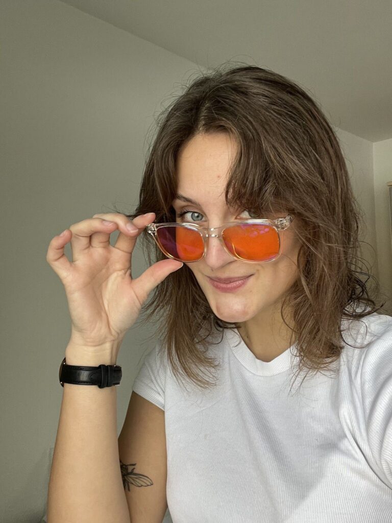 Person mit braunen Haaren und weißem Shirt hält lächelnd orange-verspiegelte Brille